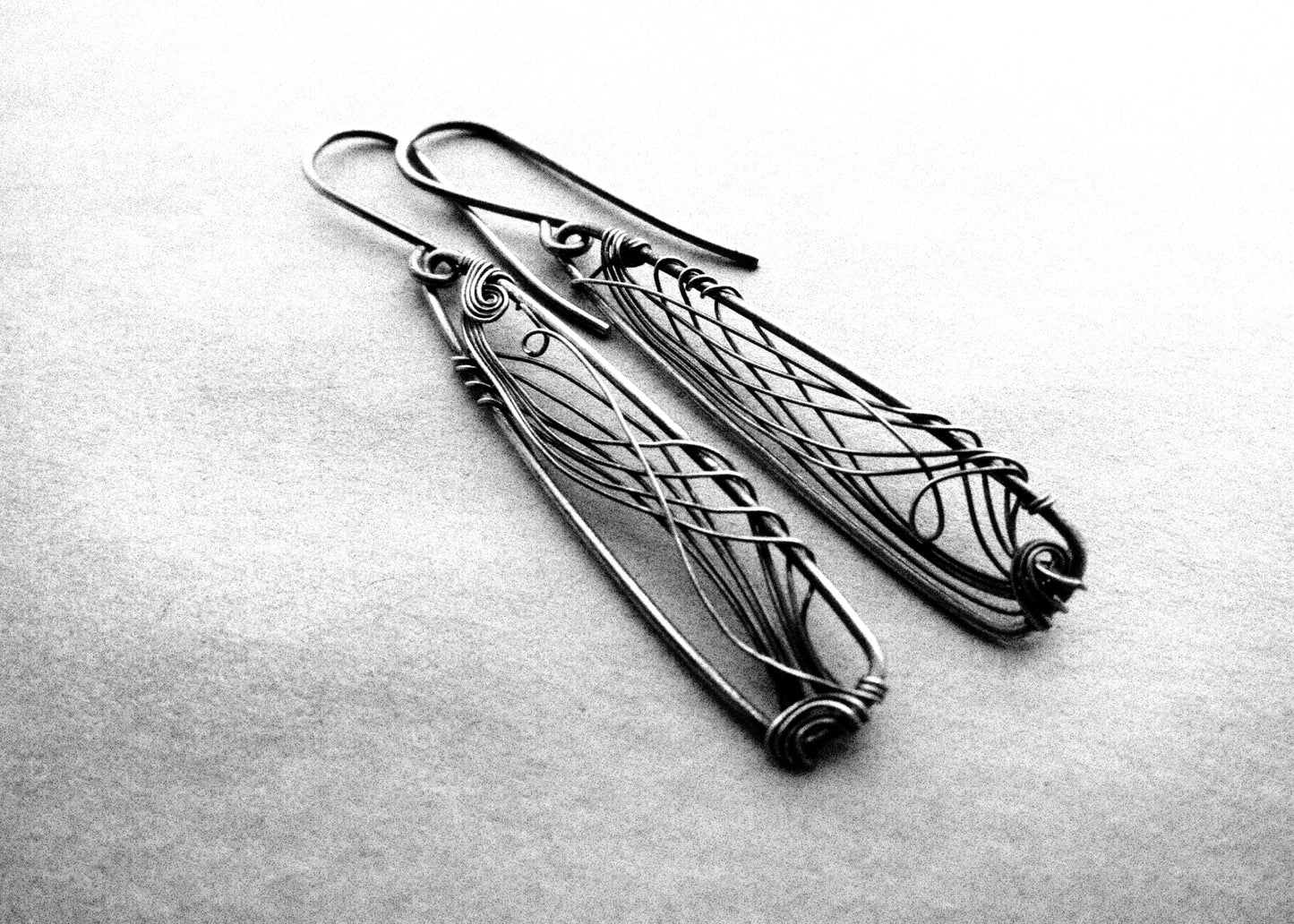 A pair of long, dark gray, triangle-shaped sterling silver earrings with intricate details and thin silver wire, oxidized and polished for a textured finish, hanging on silver ear wires.
