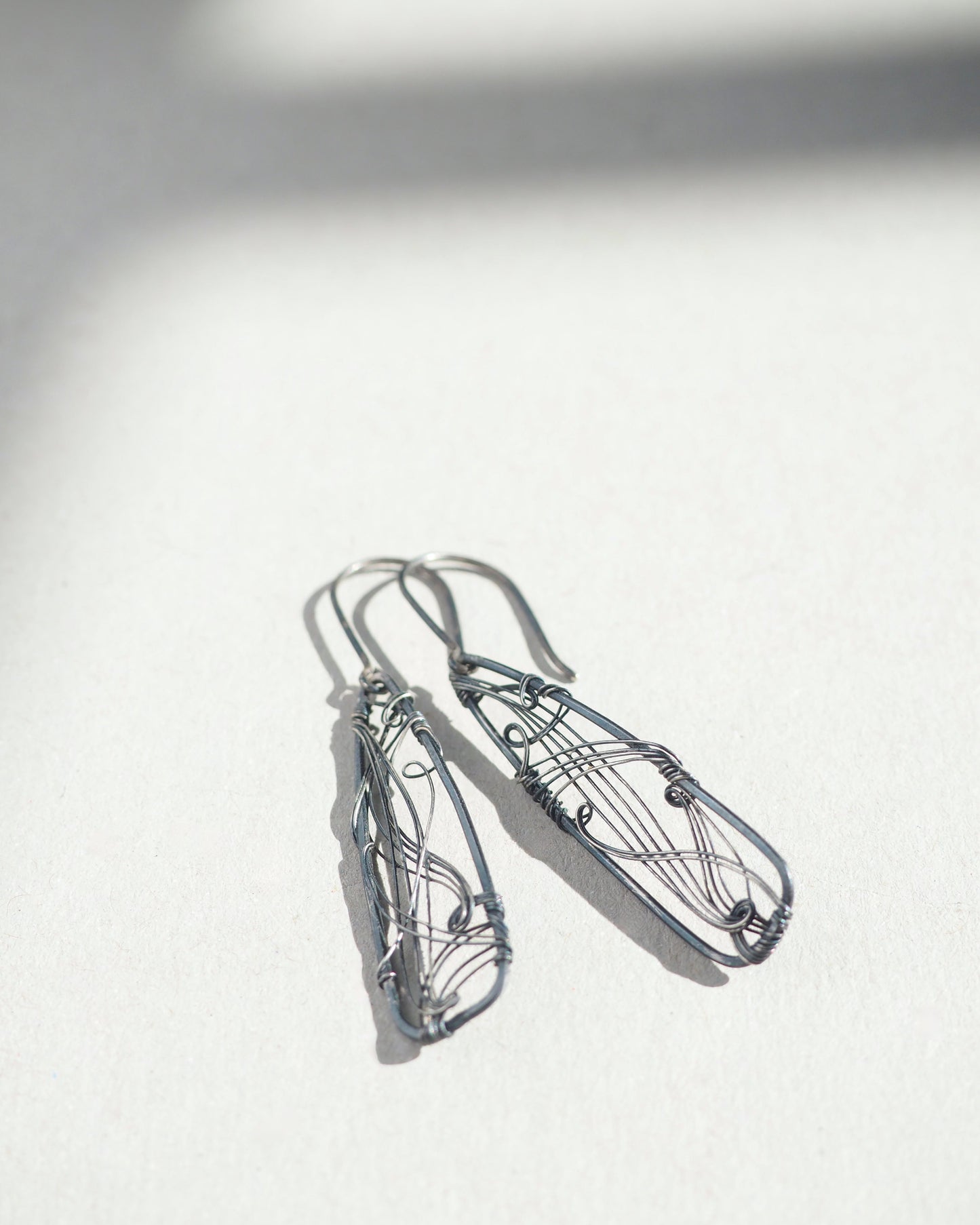 A pair of long, dark gray, triangle-shaped sterling silver earrings with intricate details and thin silver wire, oxidized and polished for a textured finish, hanging on silver ear wires.