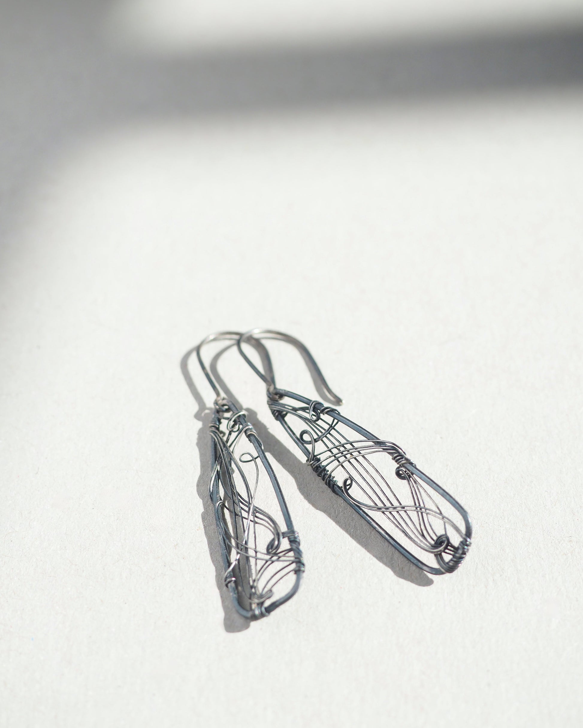 A pair of long, dark gray, triangle-shaped sterling silver earrings with intricate details and thin silver wire, oxidized and polished for a textured finish, hanging on silver ear wires.