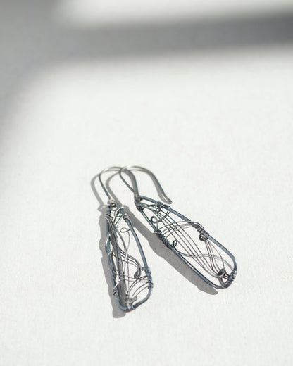 A pair of long, dark gray, triangle-shaped sterling silver earrings with intricate details and thin silver wire, oxidized and polished for a textured finish, hanging on silver ear wires.