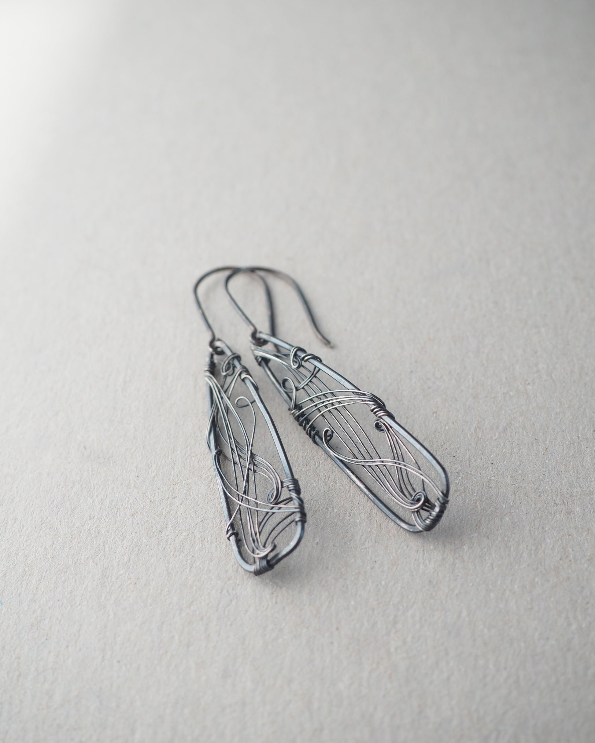 A pair of long, dark gray, triangle-shaped sterling silver earrings with intricate details and thin silver wire, oxidized and polished for a textured finish, hanging on silver ear wires.