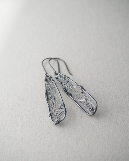A pair of long, dark gray, triangle-shaped sterling silver earrings with intricate details and thin silver wire, oxidized and polished for a textured finish, hanging on silver ear wires.