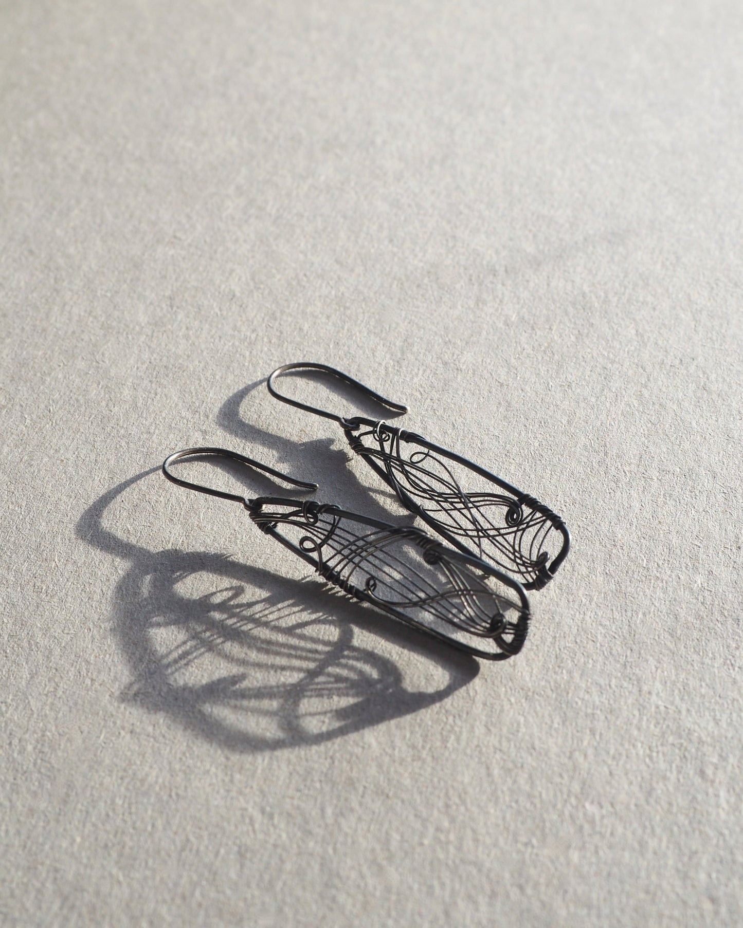 A pair of long, dark gray, triangle-shaped sterling silver earrings with intricate details and thin silver wire, oxidized and polished for a textured finish, hanging on silver ear wires.