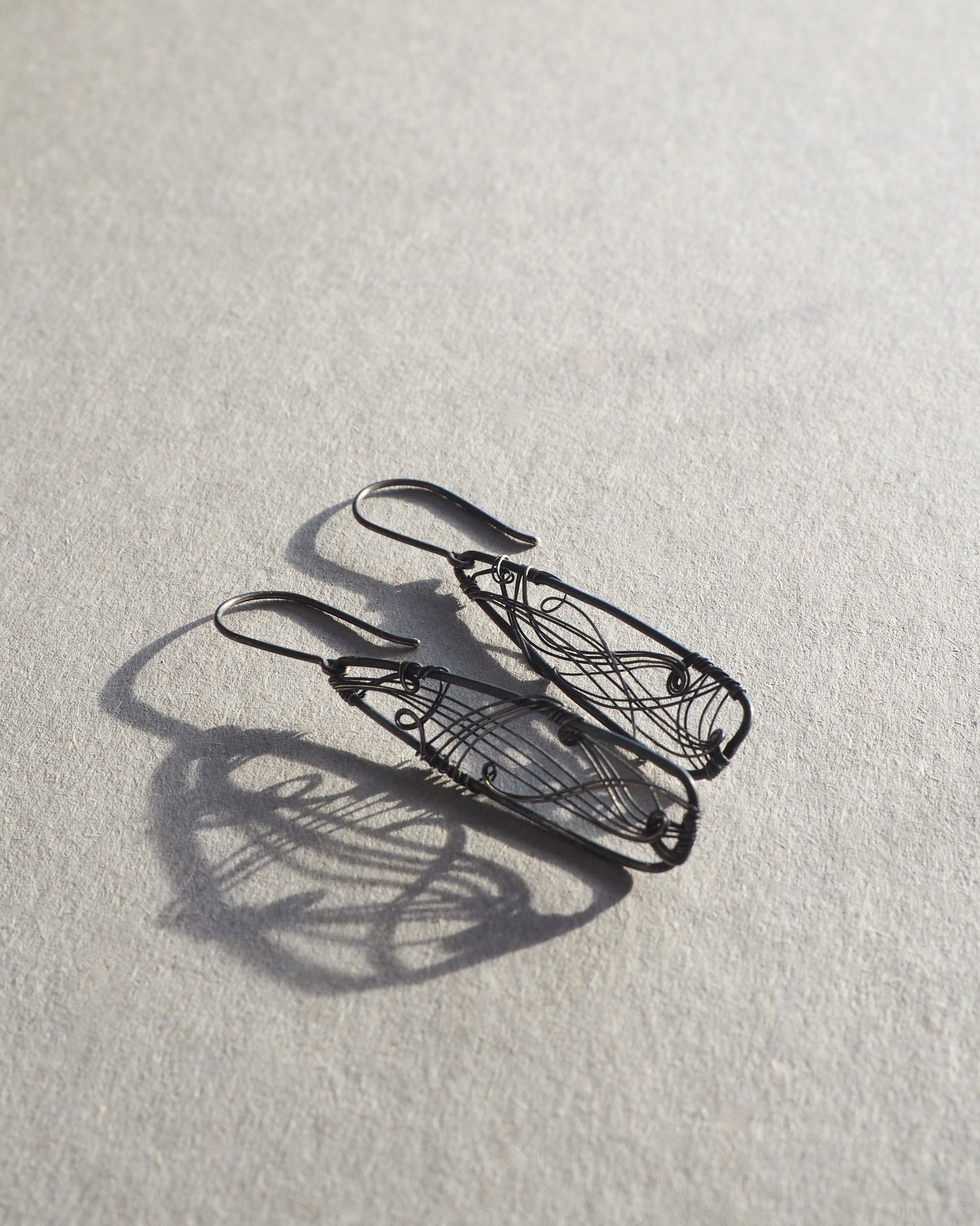 A pair of long, dark gray, triangle-shaped sterling silver earrings with intricate details and thin silver wire, oxidized and polished for a textured finish, hanging on silver ear wires.