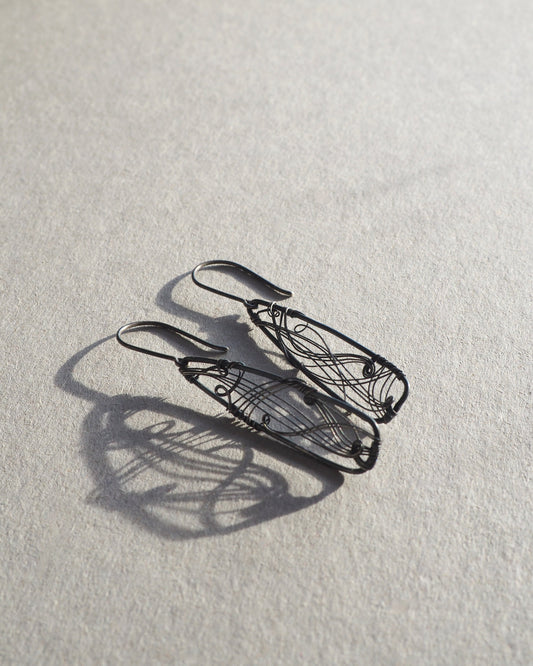 A pair of long, dark gray, triangle-shaped sterling silver earrings with intricate details and thin silver wire, oxidized and polished for a textured finish, hanging on silver ear wires.